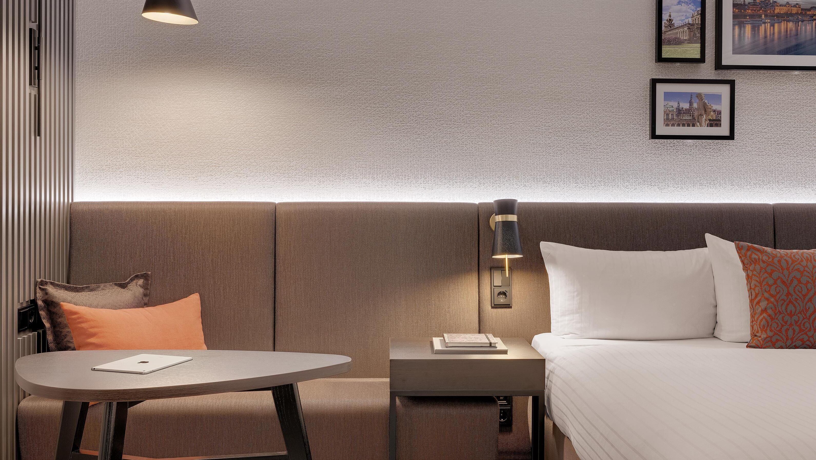 Ein Hotelzimmer mit strukturierter Wand, einem Bett mit weißen und gemusterten Kissen, einem Nachttisch mit Lampe und einem kleinen runden Tisch mit Notizbuch.