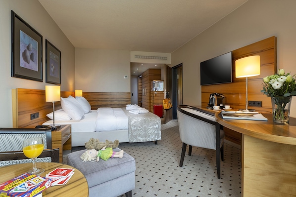 A hotel room with a bed, a desk, a chair, a television, a lamp, and a small table with a glass of orange juice and a teddy bear.