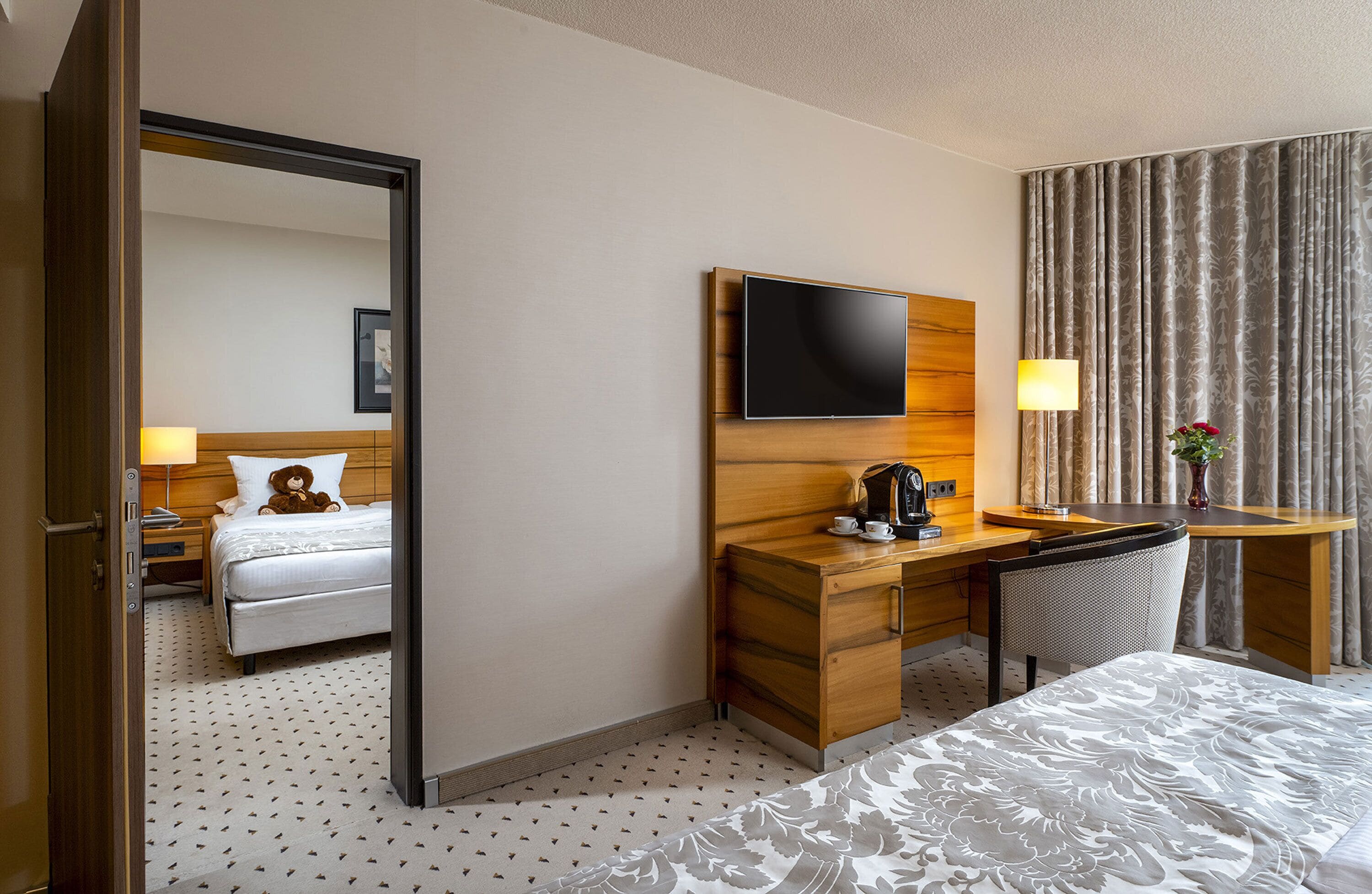 A hotel room with a bed, a desk with a flat-screen TV, a chair, a lamp, and a vase with flowers.