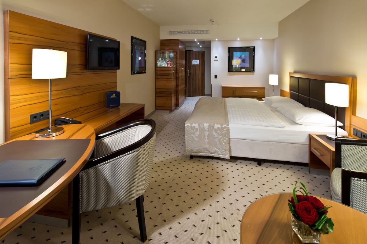 A hotel room with a bed, desk, chair, TV, wardrobe, and a vase with flowers.
