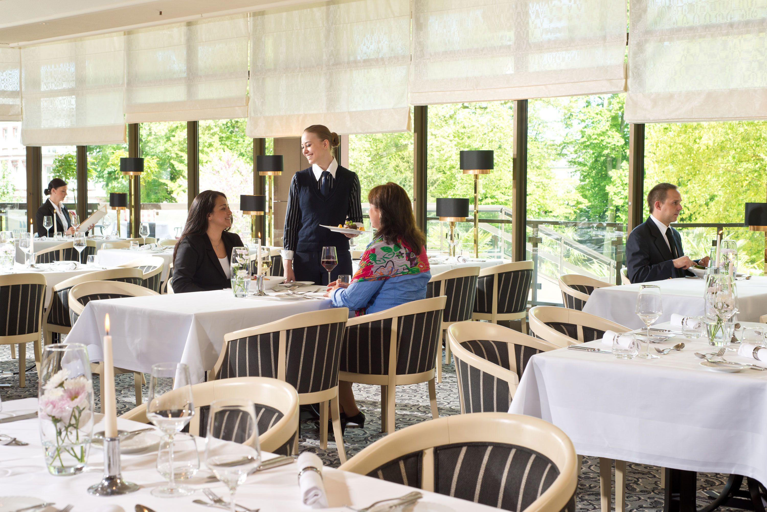 En restaurant med dækkede borde, en tjener der betjener gæster, og store vinduer med udsigt til grønne omgivelser.
