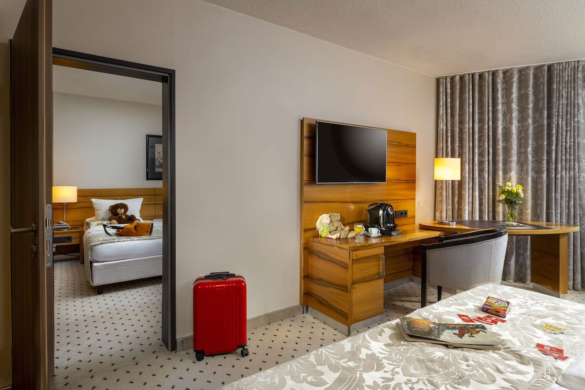 A hotel room with a bed, a desk with a TV, a chair, a red suitcase, and a small table with a magazine.