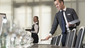 Un comedor de hotel con una larga mesa dispuesta para los huéspedes, un camarero sosteniendo una bandeja con vasos, y un hombre con traje a rayas sosteniendo un menú.