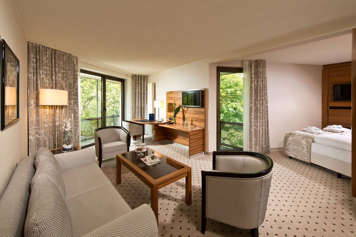 A hotel room with a sofa, a desk, a chair, a bed, and large windows offering a view of greenery.
