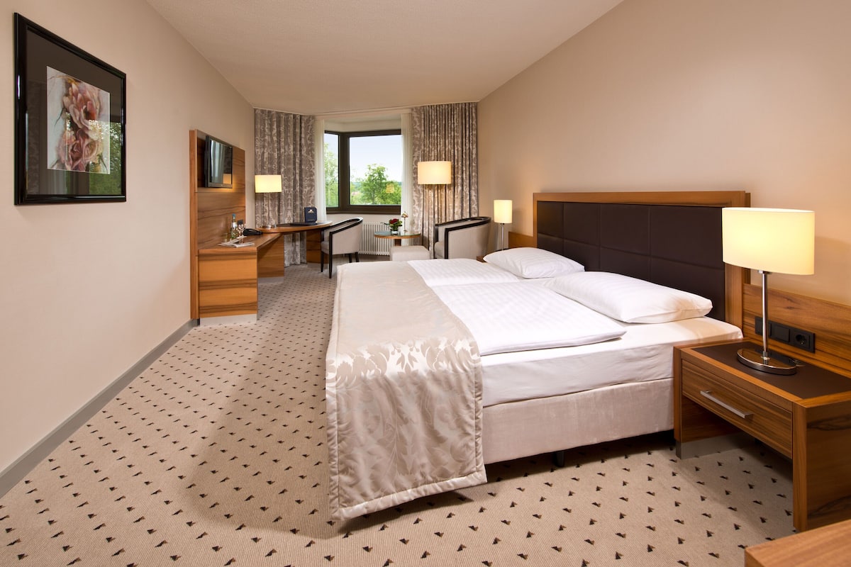 A hotel room with a bed, bedside table, desk, chair, and a window with curtains.