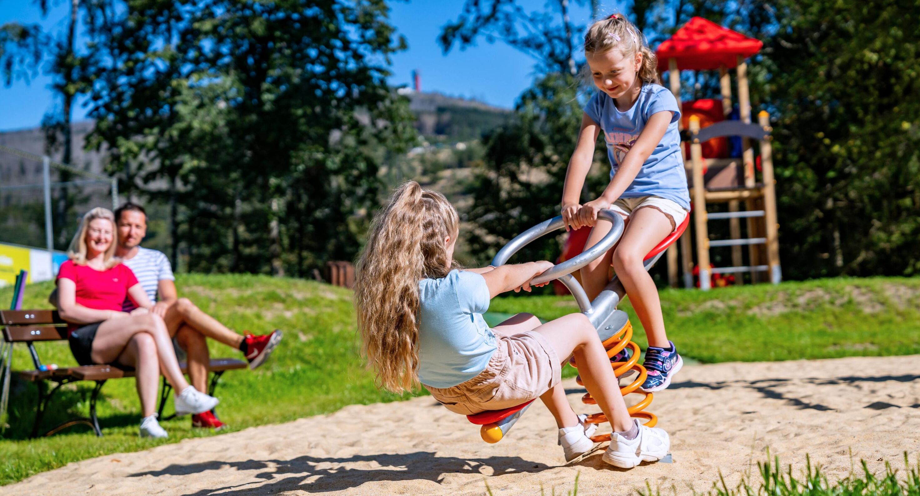 Un terrain de jeux avec une balançoire, une structure de jeu rouge et une personne assise sur un banc à l’arrière-plan.