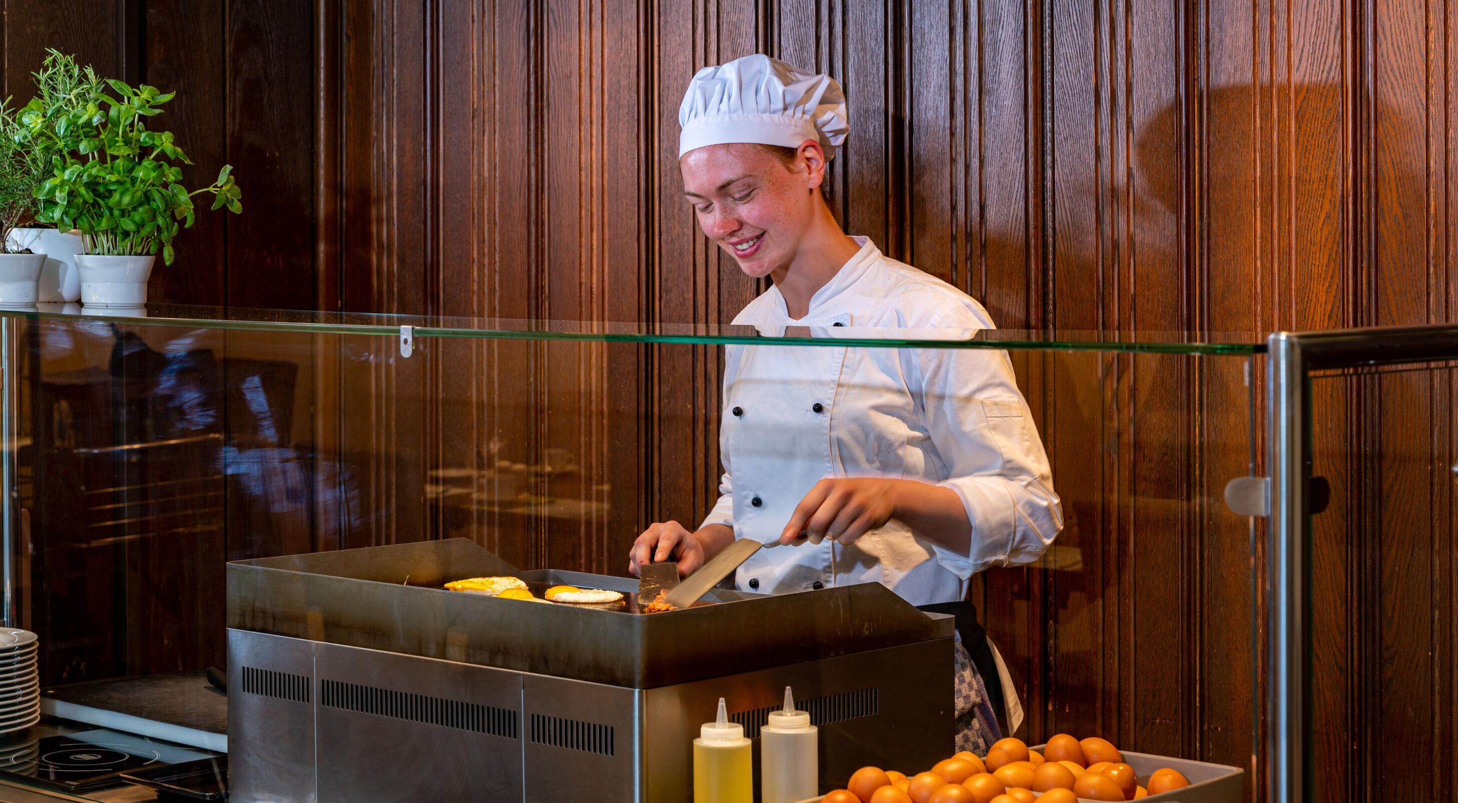 Un chef con uniforme y gorro blanco, preparando comida detrás de una mampara de vidrio en una cocina con paneles de madera.