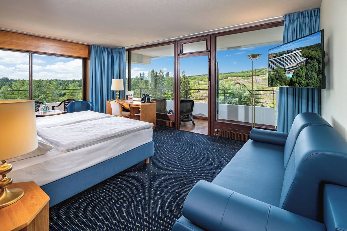 A hotel room with a bed, a sofa, a desk, and a chair. There is a large window with a view of trees and a building.