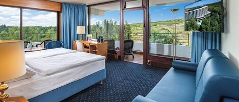 Habitación de hotel con cama, sofá, escritorio y silla. Amplia ventana con vista a árboles y un edificio.