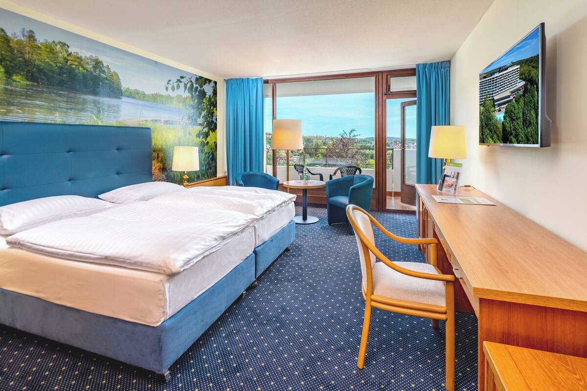 A hotel room with a large bed, a desk, two chairs, a small table, and a view of a lake through the window.