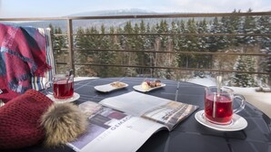 Une table avec un verre d’une boisson rouge, une assiette de desserts, un magazine et un article tricoté rouge sur un balcon donnant sur la montagne.