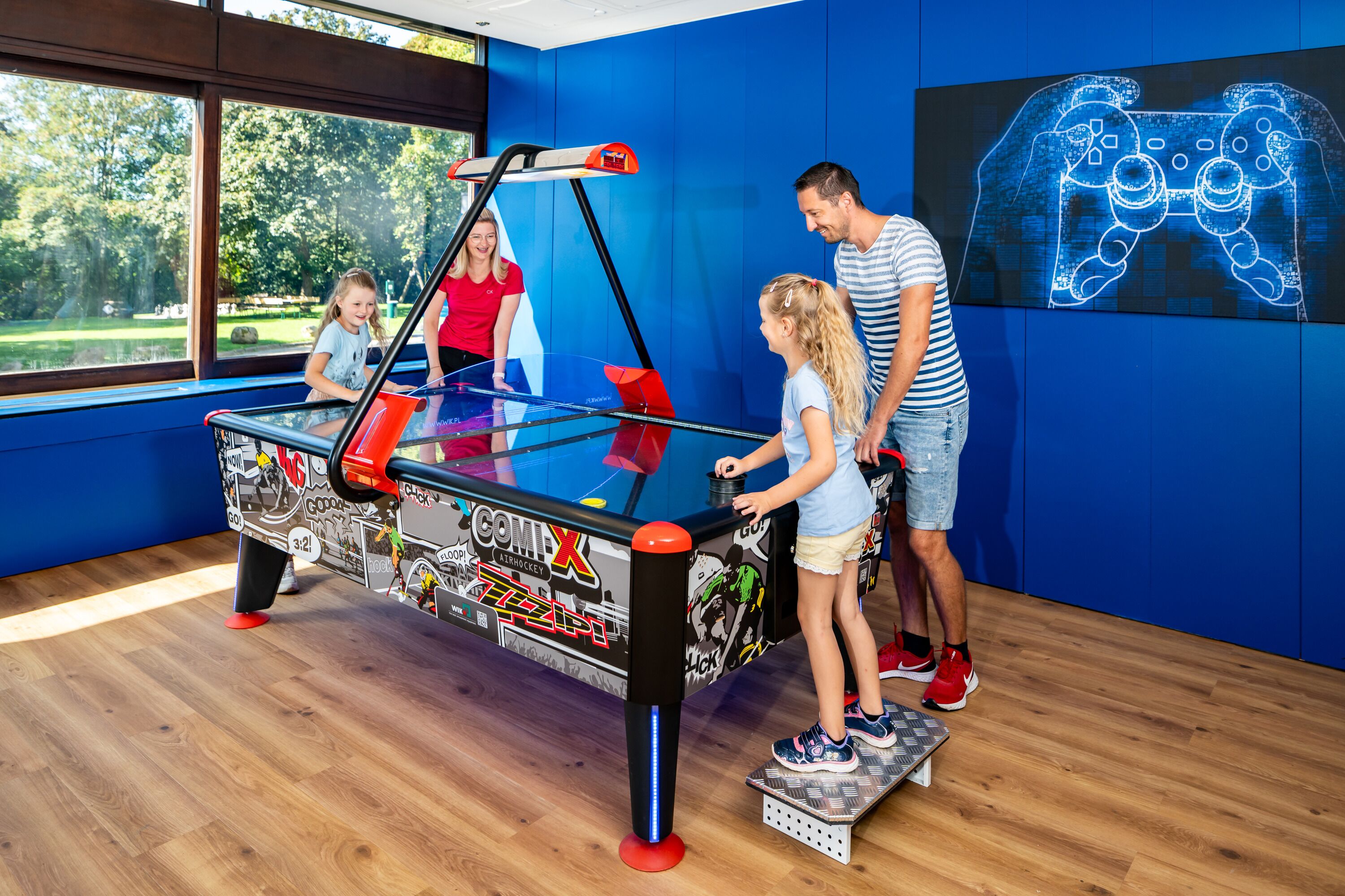 A pool table with a comic book theme, a large window, and a neon game controller artwork on the wall.
