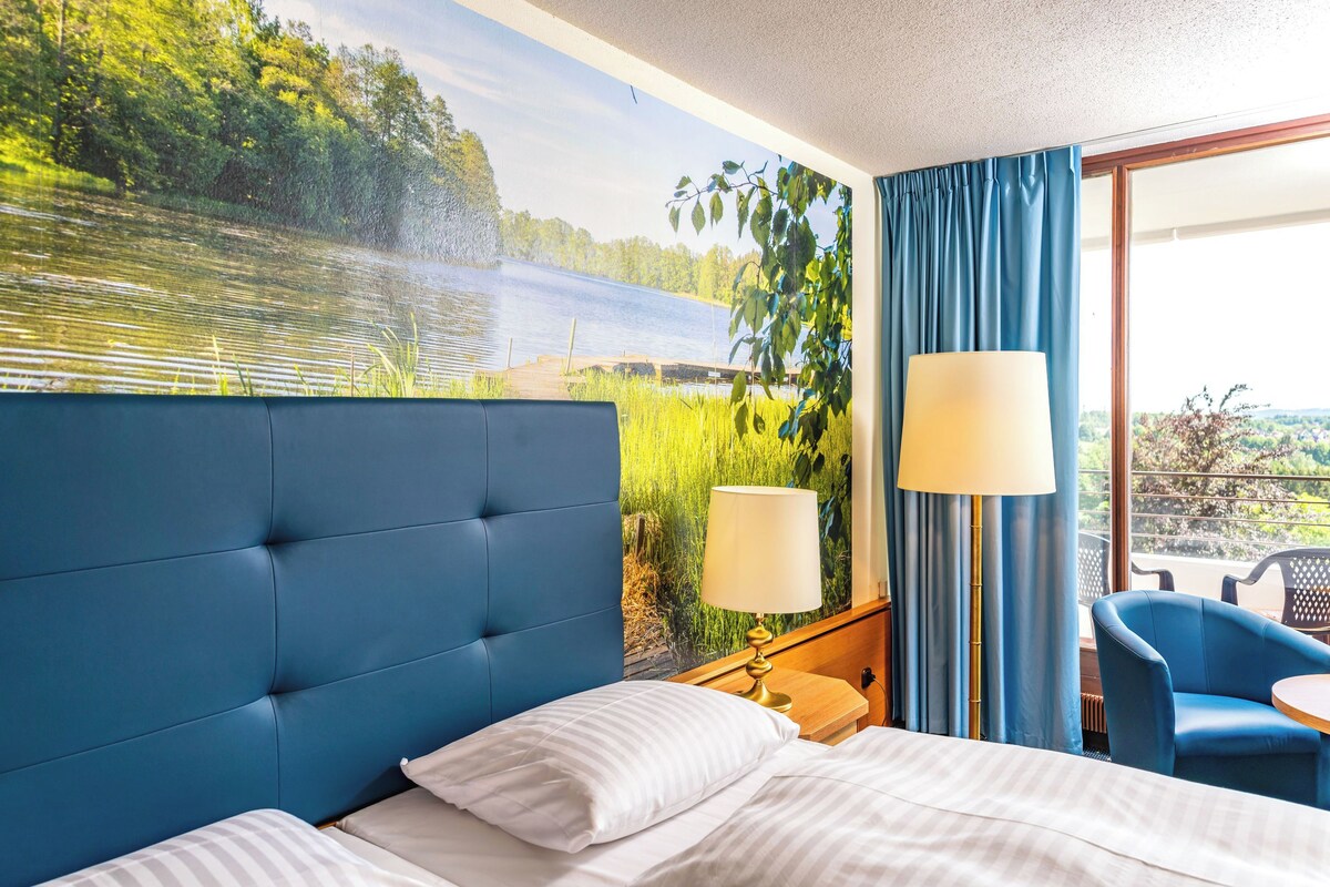 A hotel room with a large bed, a blue headboard, a window with a view of a lake, a blue chair, and a lamp.
