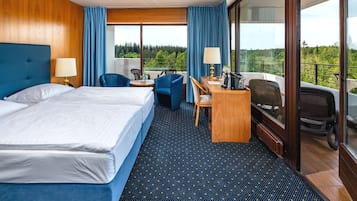 A hotel room with a large bed, a desk with a chair, a laptop, and a view of the forest.