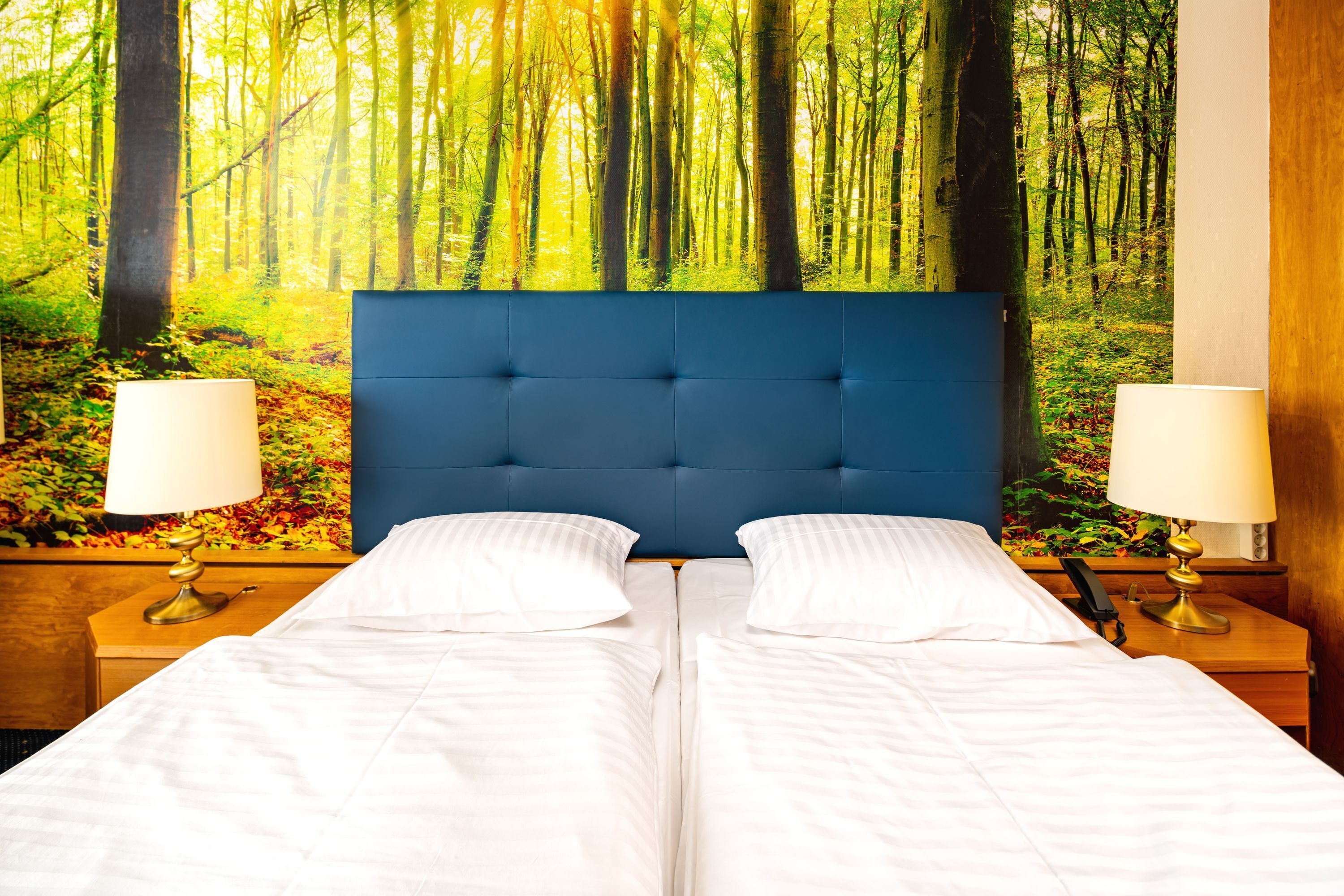 A double bed with a blue headboard, two bedside tables with lamps, and a forest mural on the wall.