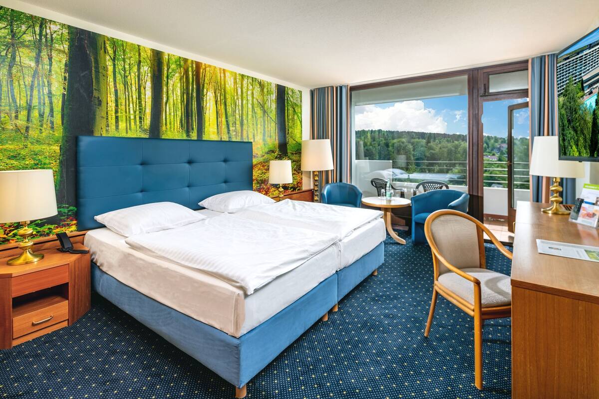 A hotel room with a large bed, two bedside tables with lamps, a desk with a chair, and a view of a forest through a large window.