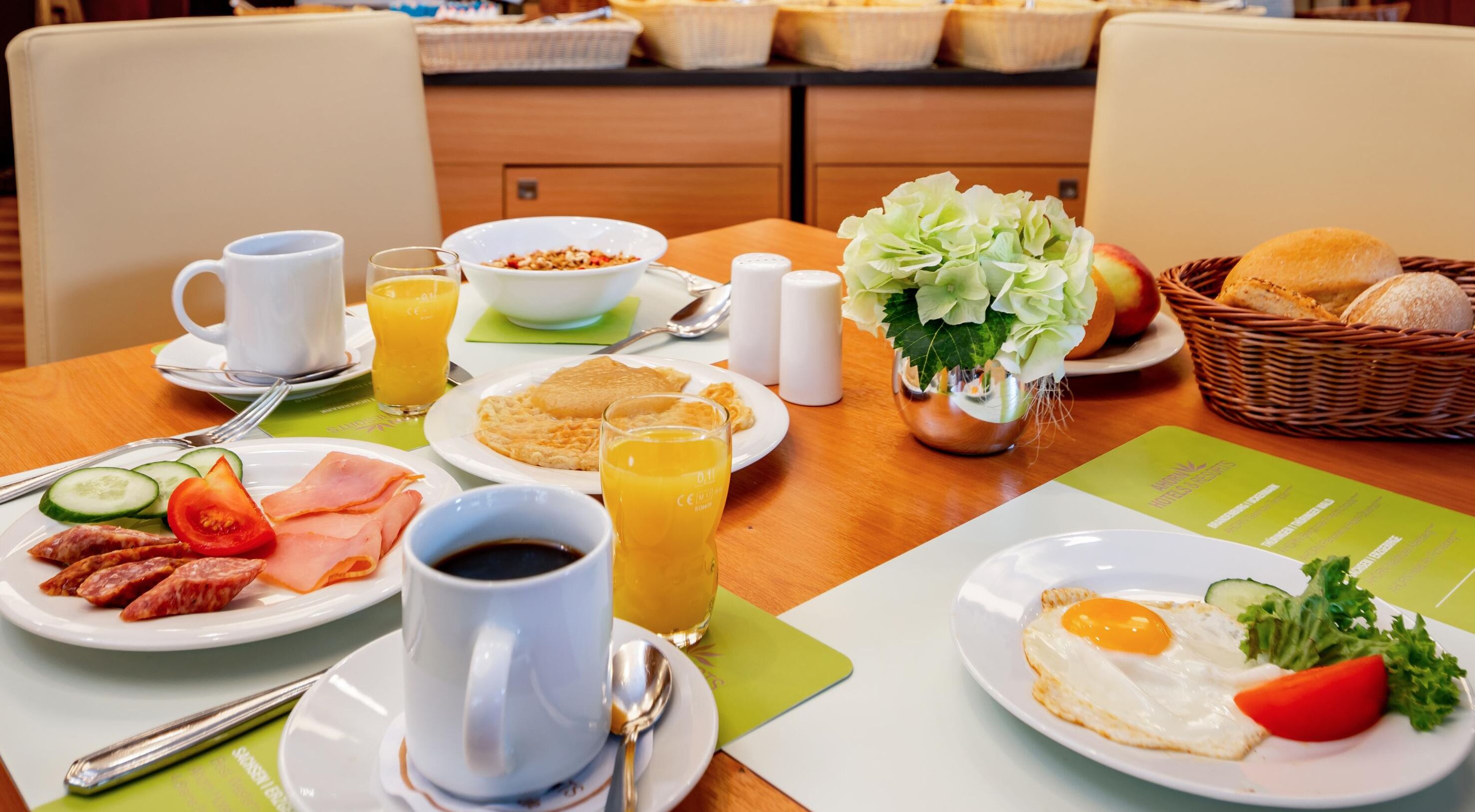 Een ontbijttafel met borden met eieren, ham en groenten, een koffiekop en een glas sinaasappelsap.