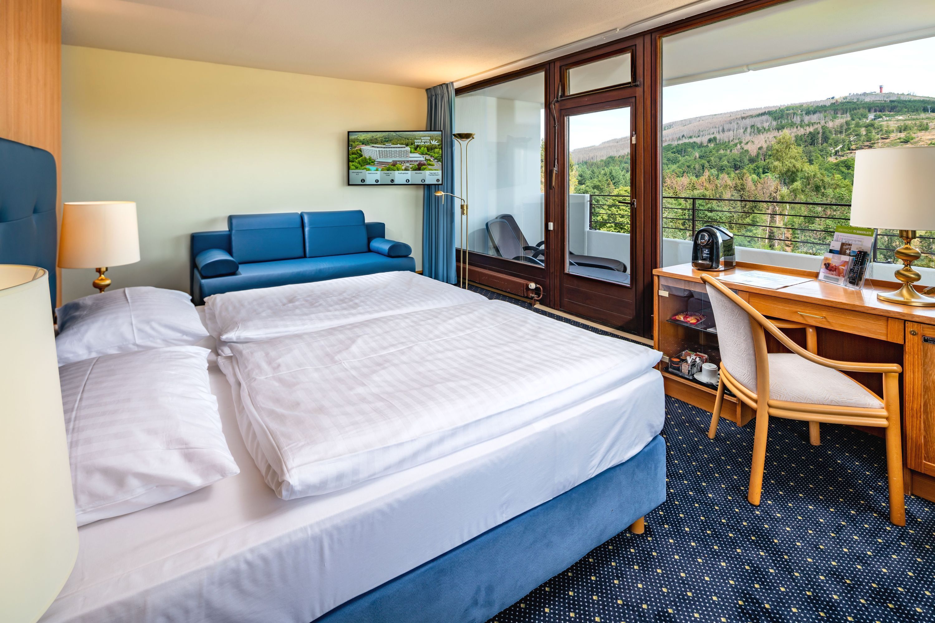 Habitación de hotel con una cama grande, un escritorio con silla, un sofá azul y vistas a un paisaje montañoso desde un balcón.