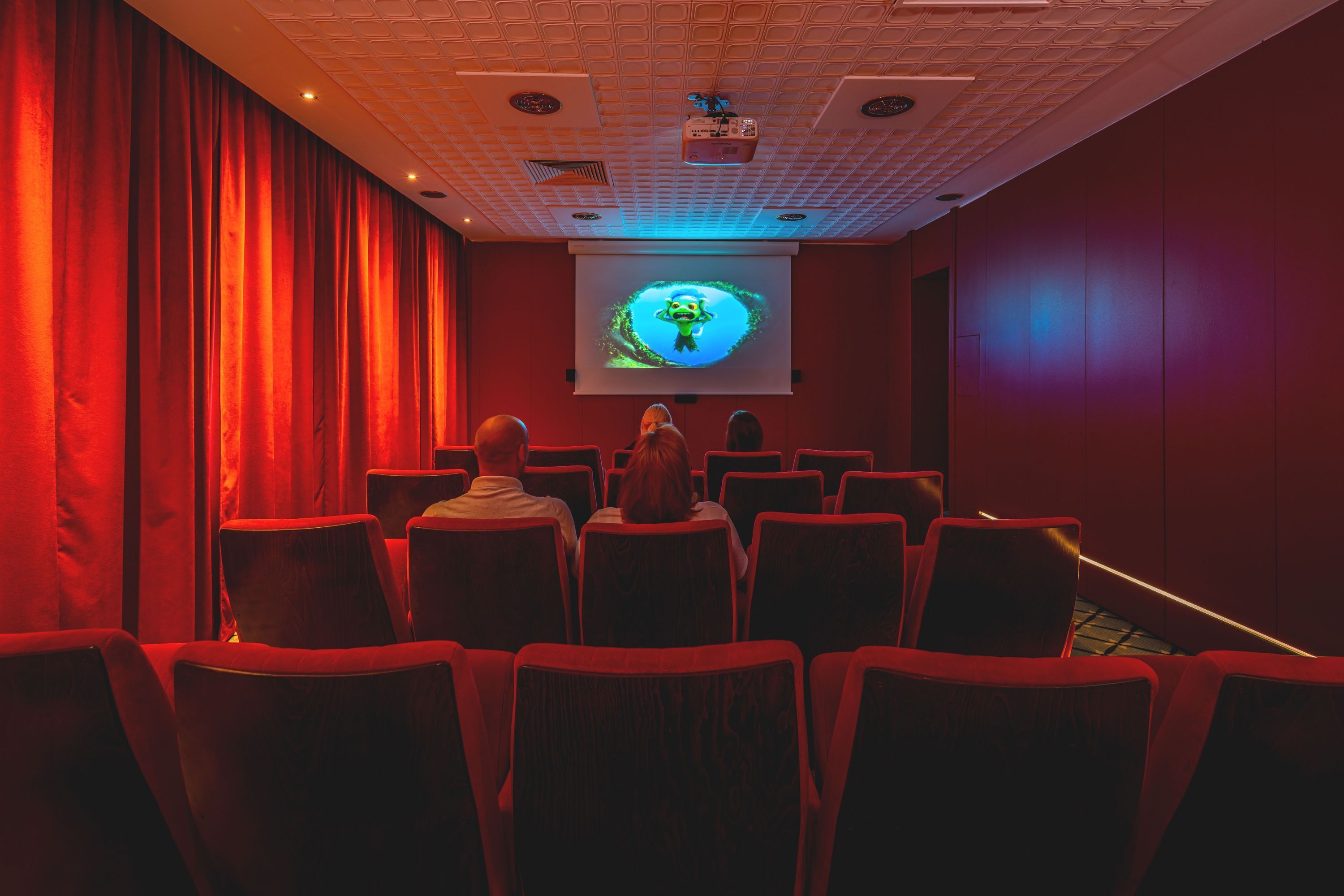 A theater with red curtains, rows of seats, and a screen displaying a green animated character.