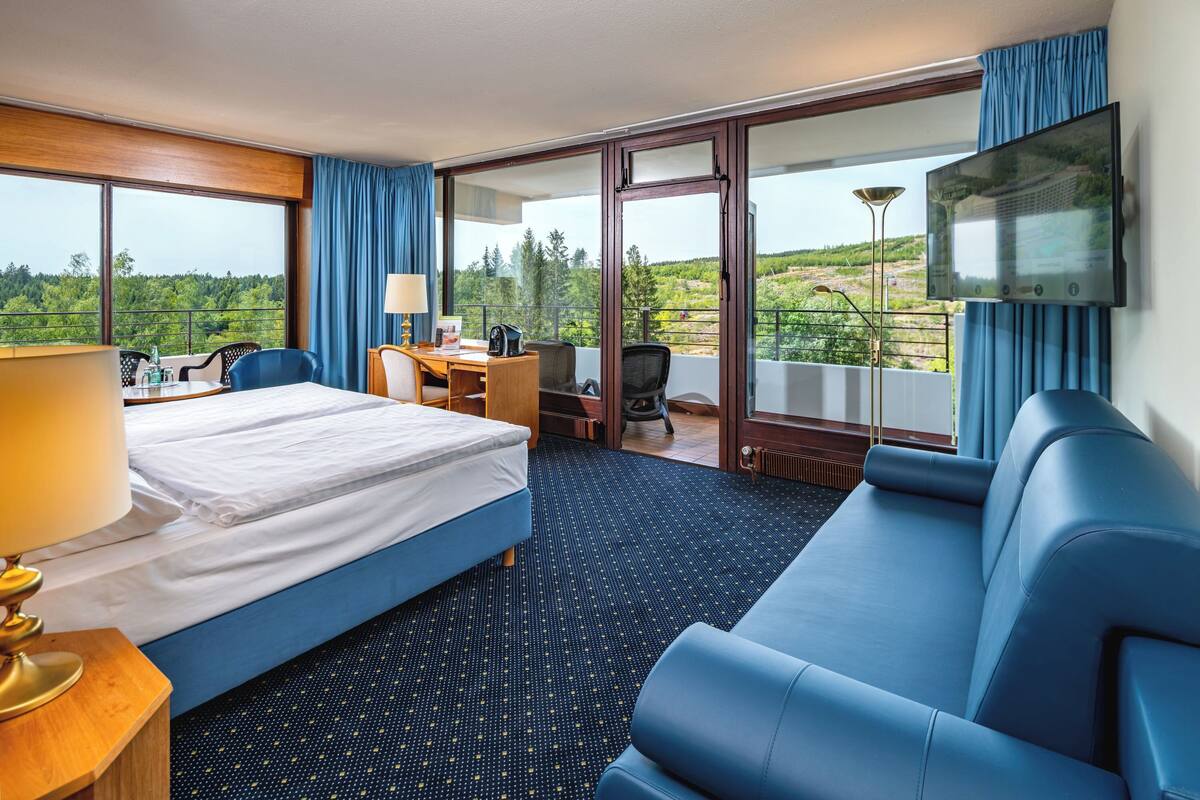 A hotel room with a bed, a desk, a chair, a sofa, a TV, and a balcony with a view.