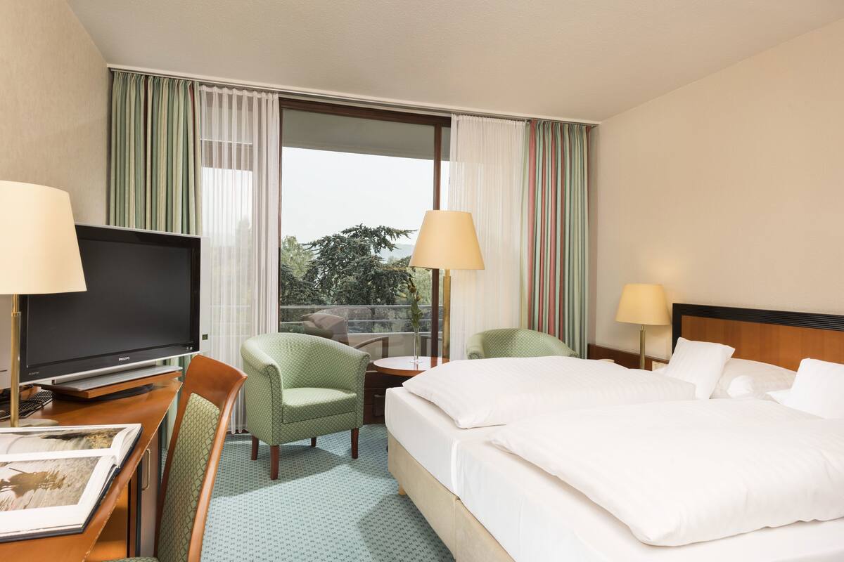 Una habitación de hotel con una cama, dos sillas, un escritorio con televisión y una ventana con cortinas.