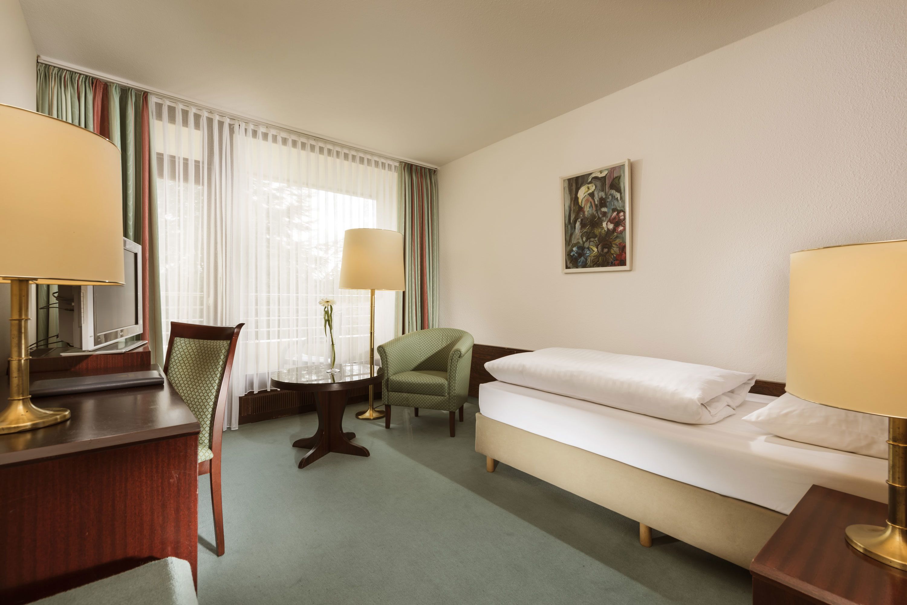 A hotel room with a bed, a desk, a chair, a small table, and a lamp.