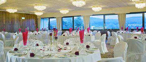 Un salón de banquetes con mesas redondas dispuestas para un evento formal, grandes ventanales con vistas a la montaña y candelabros.