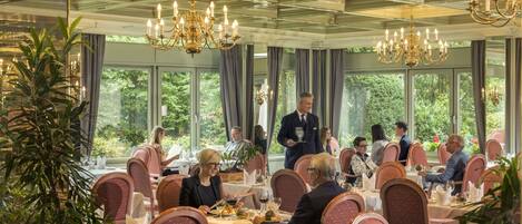 Un comedor elegante con mesas dispuestas para invitados, ventanas amplias con cortinas y candelabros.