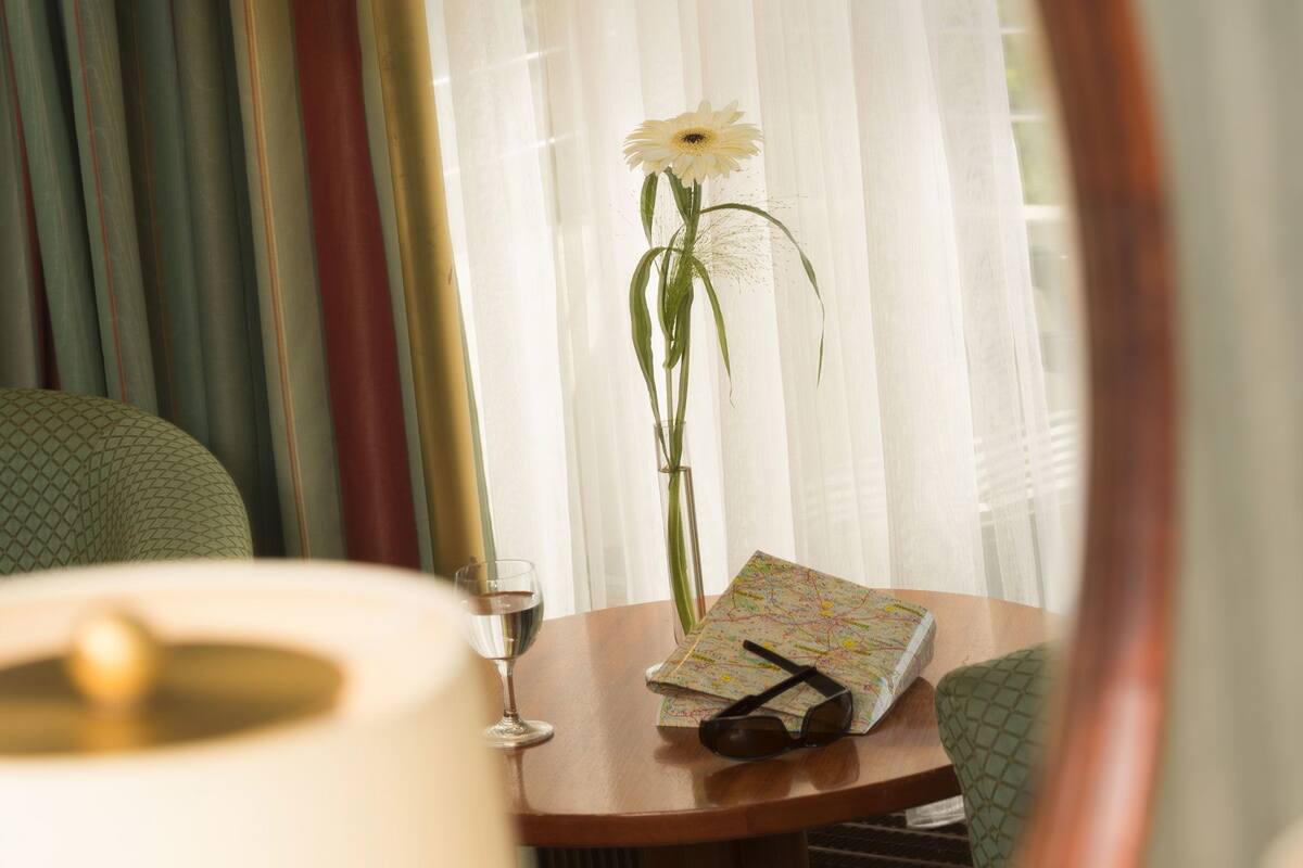 Una habitación con un espejo que refleja una lámpara, un florero con una flor, una copa de vino y un mapa sobre una mesa.