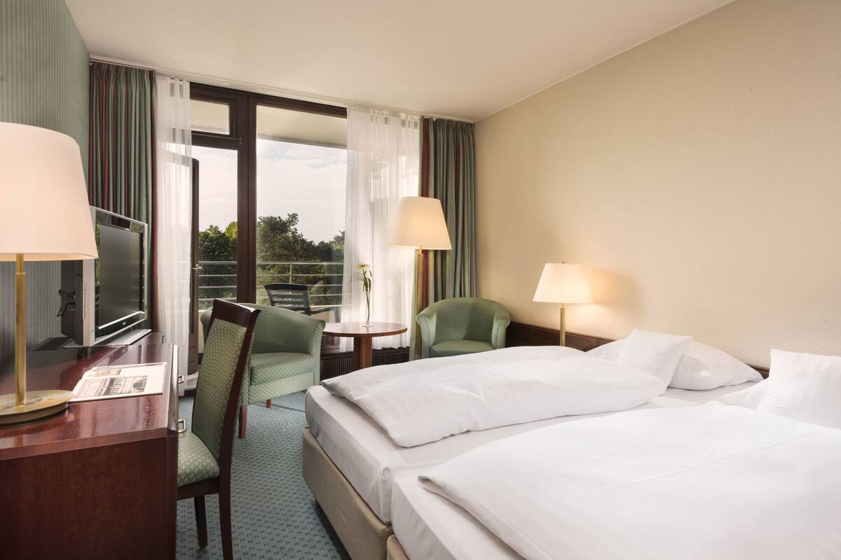 Habitación de hotel con cama, dos sillones, un escritorio con lámpara, televisión y ventana con cortinas.