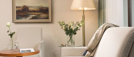 A hotel room with a sofa, a small table with a vase of flowers, a lamp, and a framed painting on the wall.