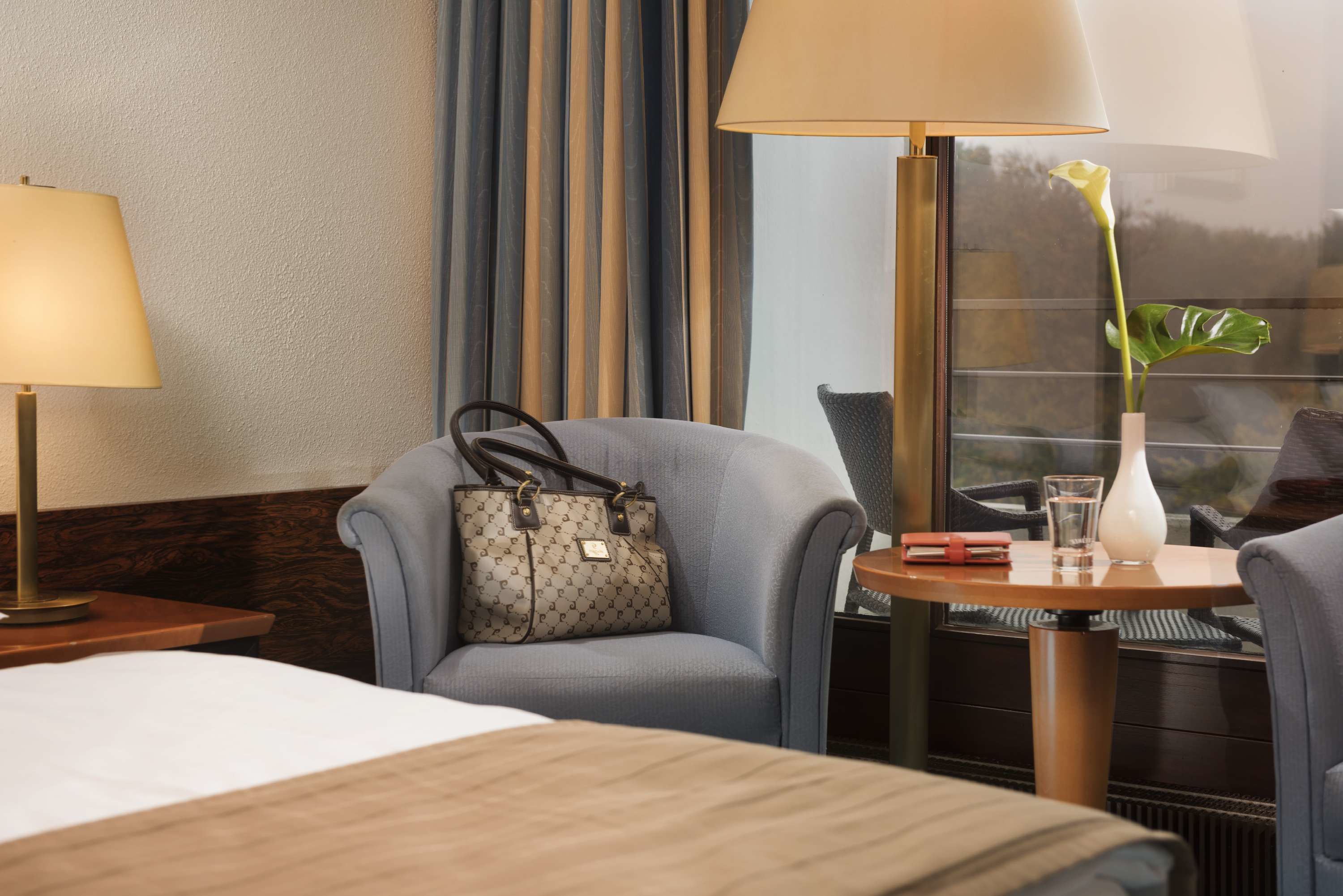 Habitación de hotel con una cama, una silla, una mesa con un jarrón y un vaso, y una lámpara.