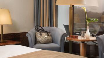 A hotel room with a bed, a chair, a table with a vase and a glass, and a lamp.
