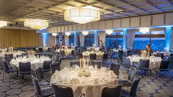 Een balzaal met ronde tafels, gedekt voor een formeel evenement, met kroonluchters en uitzicht op de stad.