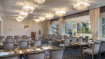 Un salón de conferencias con varias mesas y sillas, ventanas amplias con cortinas y candelabros.