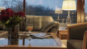Een kamer met een glazen tafel, een vaas rode rozen, een drankje, een tijdschrift, een lamp en uitzicht op bomen door een raam.