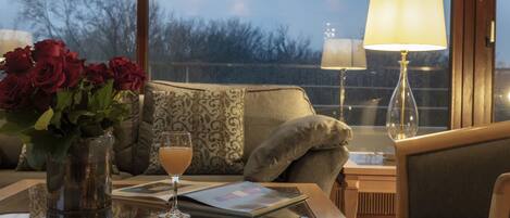 A room with a glass table, a vase of red roses, a glass of drink, a magazine, a lamp, and a view of trees through a window.