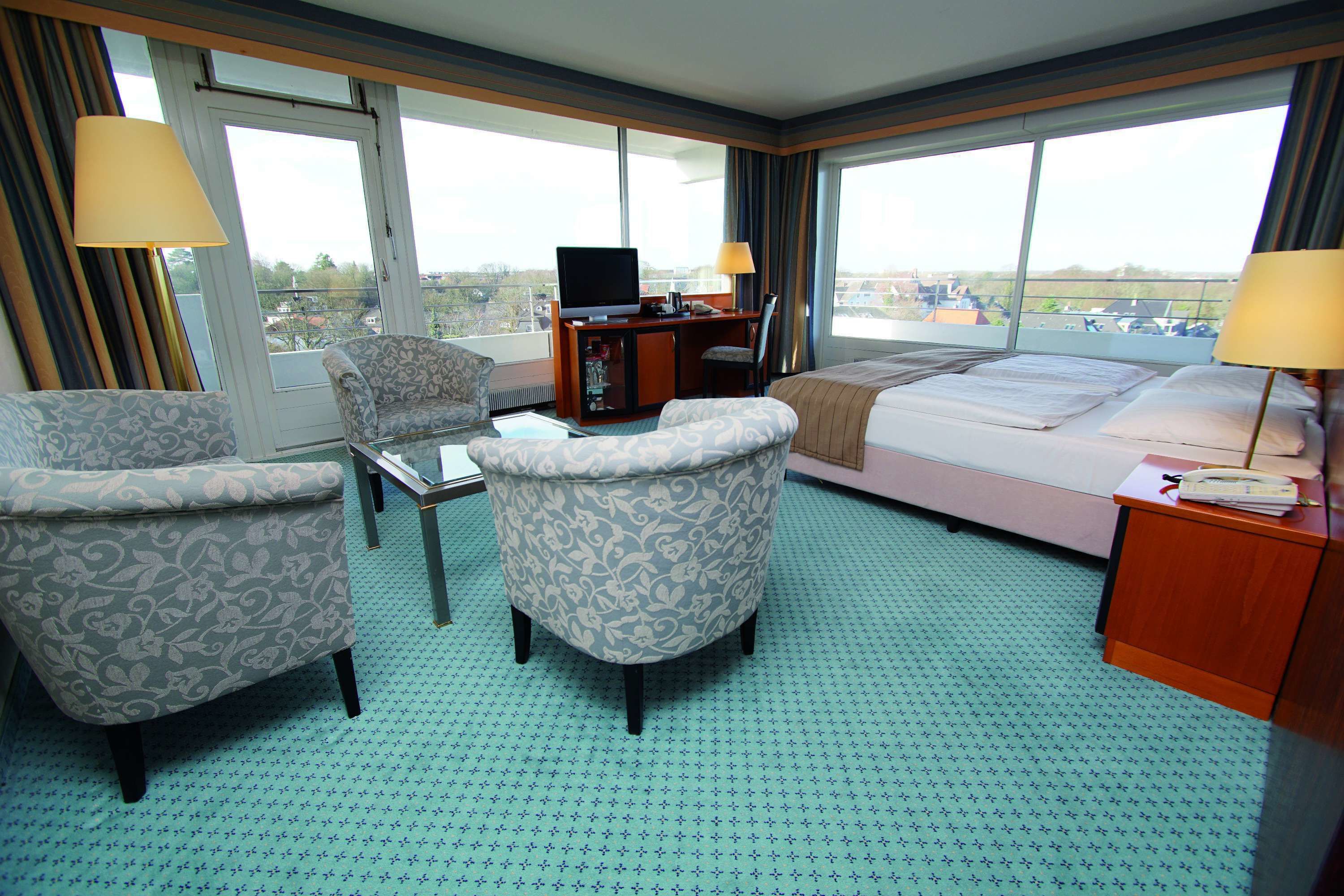 A hotel room with a bed, two armchairs, a desk with a TV, and large windows offering a city view.