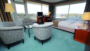 A hotel room with a bed, two armchairs, a desk with a TV, and large windows offering a city view.