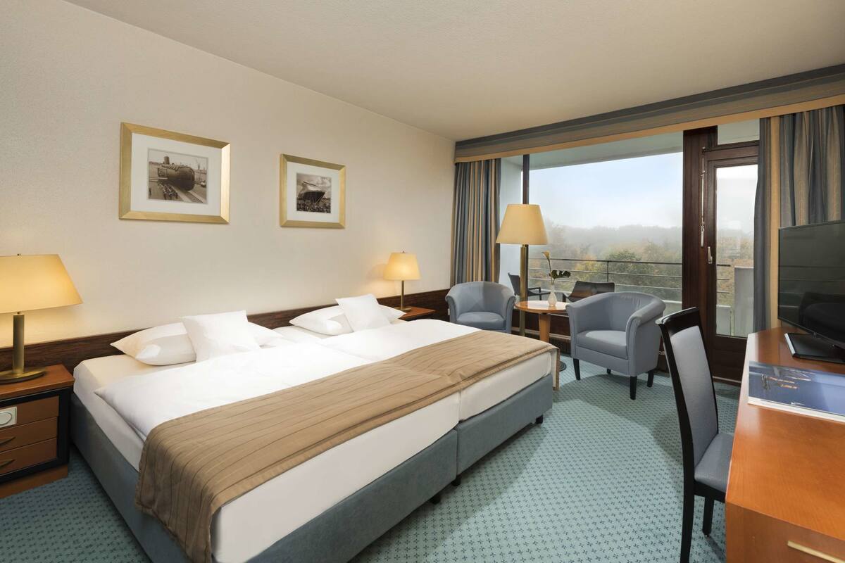Ein Hotelzimmer mit einem großen Bett, zwei Nachttischlampen, einem Schreibtisch mit Stuhl, einem Fernseher und einem Fenster mit Vorhängen.