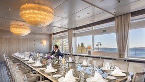 Een feestzaal met een lange tafel gedekt voor een formeel evenement, grote ramen met uitzicht en elegante kroonluchters.