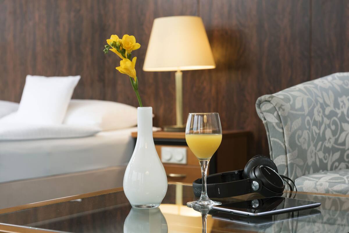 Ein Hotelzimmer mit einem Bett, einem Nachttisch mit Lampe, einer Vase mit Blumen, einem Glas Orangensaft und Kopfhörern auf dem Tisch.