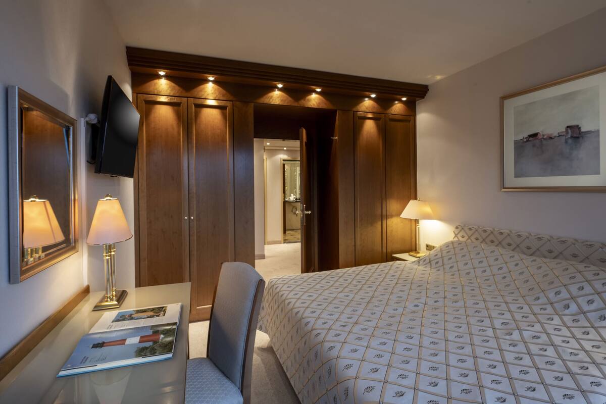 Ein Hotelzimmer mit Bett, Schreibtisch mit Lampe, Sessel und einem an der Wand befestigten Fernseher sowie einem gerahmten Bild.