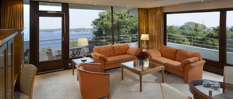 A modern living room with a sofa set, a coffee table, and a glass table. There is a view of a body of water through large windows.