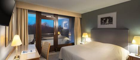 A hotel room with a large bed, two bedside tables with lamps, a desk with a chair, a large window with curtains, and a framed picture on the wall.