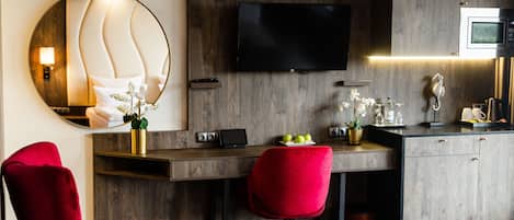 A modern hotel room with a wooden desk, a flat-screen TV, a red chair, and a mirror with a light fixture.