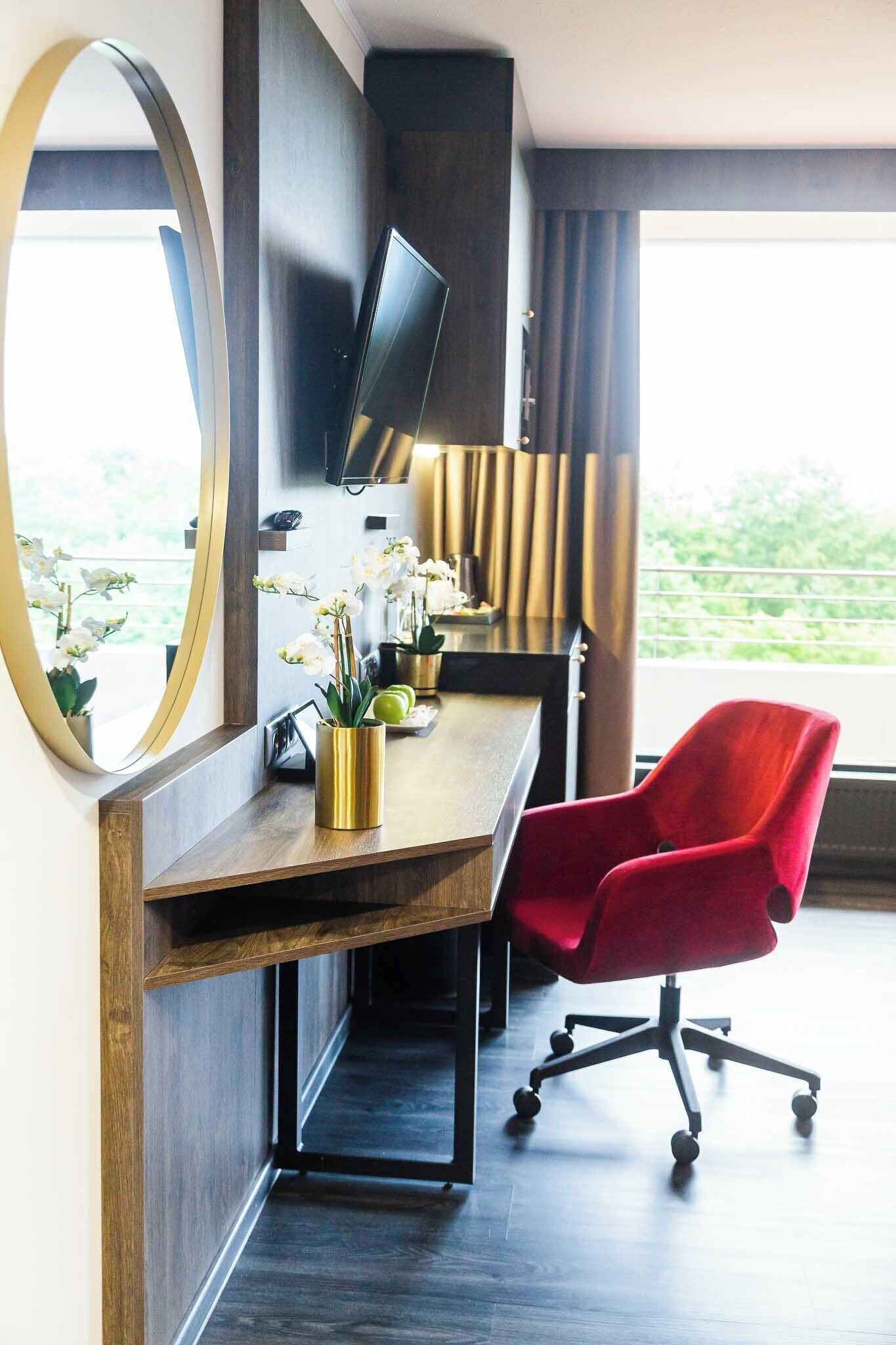 A modern office space with a desk, a red office chair, a television mounted on the wall, and a view of greenery outside.