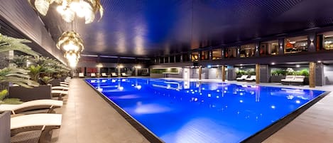 A large indoor swimming pool with a modern design, surrounded by lounge areas and a ceiling with a unique pattern.
