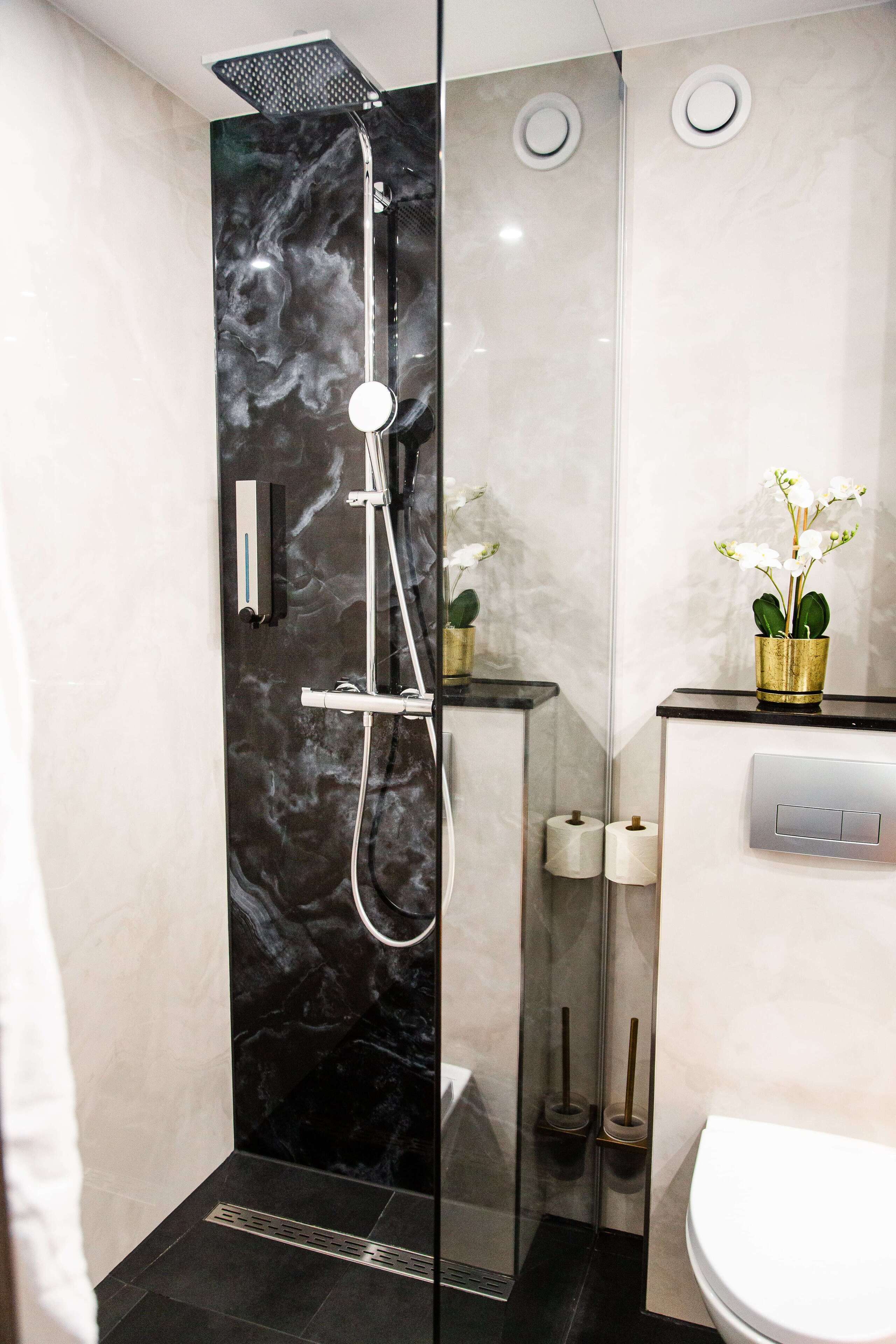 Un baño moderno con ducha envuelta en cristal, paredes de mármol, un inodoro y una pequeña orquídea en maceta sobre el mueble.