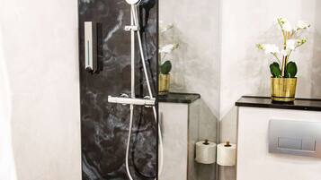 Un baño moderno con ducha envuelta en cristal, paredes de mármol, un inodoro y una pequeña orquídea en maceta sobre el mueble.